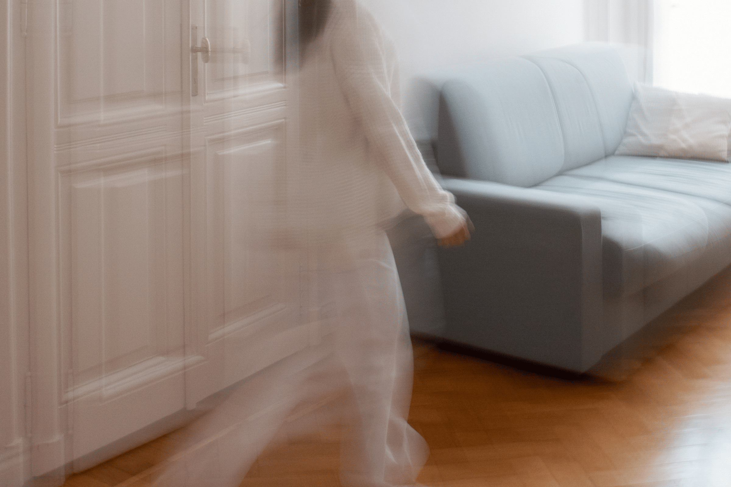Motion-blurred person in a beige coat walking past a white paneled door toward a light-blue sofa in a bright living room area.