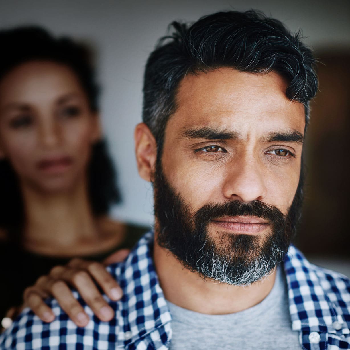 Close-up of a bearded man looking pensive, with a woman resting a hand on his shoulder in the blurred background.
