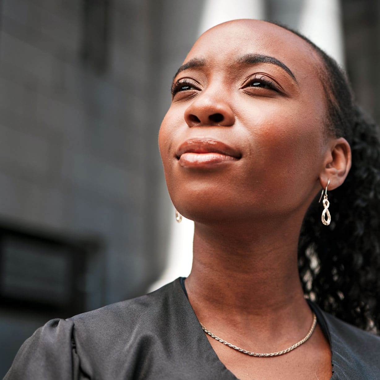 Close-up of a young Black woman looking up with a calm smile, wearing gold teardrop earrings and a gold necklace.